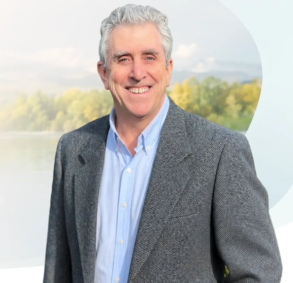 TraumaSolutions A man with gray hair wearing a gray blazer and light blue shirt smiles outdoors with trees and water in the background, embodying the calming presence of an experienced trauma therapist specializing in EFT (Emotional Freedom Techniques).