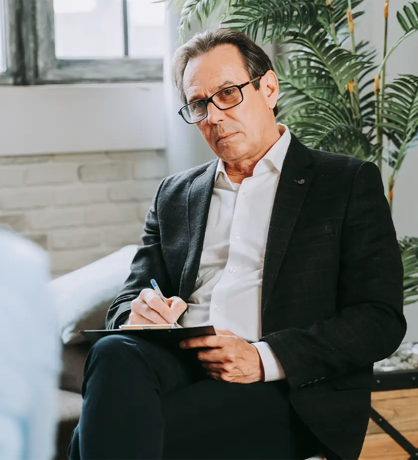 TraumaSolutions Middle-aged man in glasses and a suit sits on a chair, holding a clipboard and pen, listening attentively—embodying the calm presence of an experienced trauma therapist. Indoor setting with a plant and window in the background.