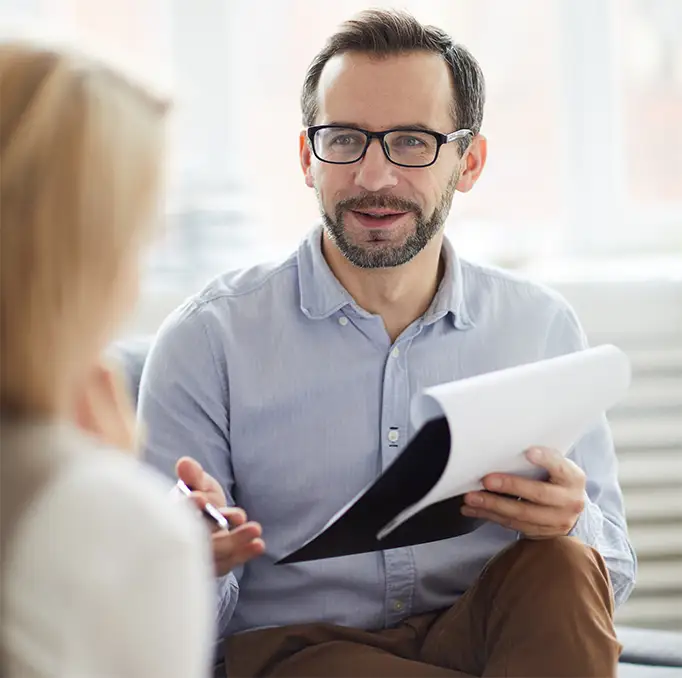 TraumaSolutions A man wearing glasses and holding papers appears to be talking to a woman in an office or meeting setting, possibly acting as a trauma therapist or EFT guide.