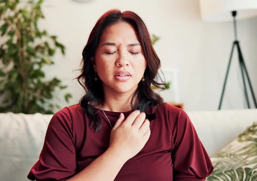 TraumaSolutions A woman sitting indoors, perhaps guided by an EFT or trauma therapist, touches her upper chest with her eyes closed, appearing to take a deep breath. Green plants and home decor are visible in the background.