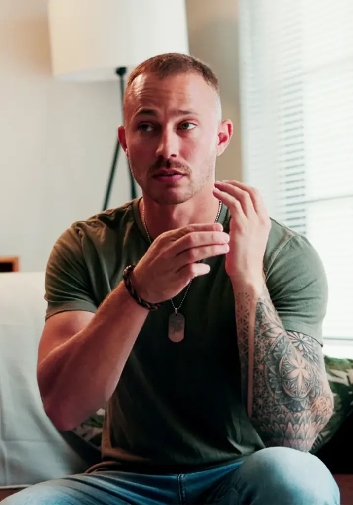 TraumaSolutions A man with a buzz cut and arm tattoo sits indoors, gesturing with his hands like an experienced trauma therapist. He wears a green t-shirt, jeans, a bracelet, and a dog tag necklace.