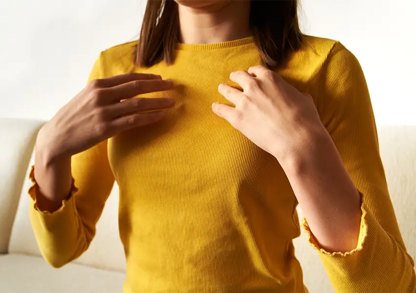 TraumaSolutions Person wearing a yellow long-sleeve shirt sits on a light-colored couch, touching their upper chest area with both hands—a gesture often seen in EFT or trauma therapy. The face is partially out of frame.