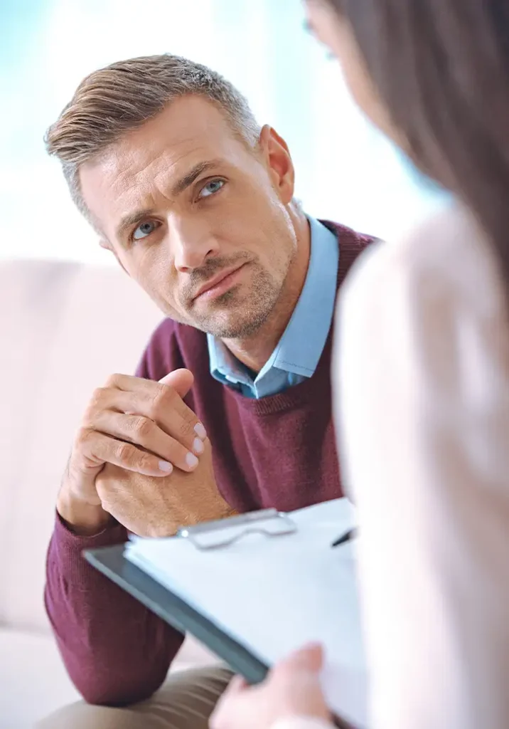 TraumaSolutions A man in a maroon sweater listens attentively to a woman holding a clipboard during a conversation, possibly in a counseling or interview setting, as she serves as his trauma therapist and guide.