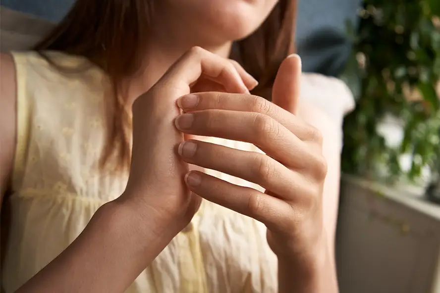 TraumaSolutions A person in a sleeveless yellow top holds and gently massages their left hand with their right hand, practicing an EFT technique. Green plants are visible in the background, adding a calming touch to this mindful self-care moment.