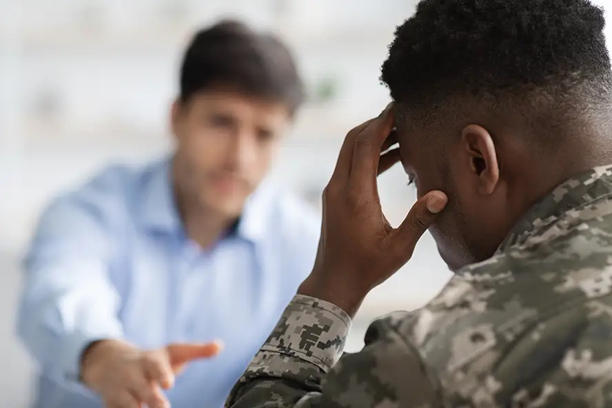 TraumaSolutions A person in military uniform holds their head in their hand while another person in a blue shirt, possibly serving as a trauma therapist or guide, gestures toward them in conversation.