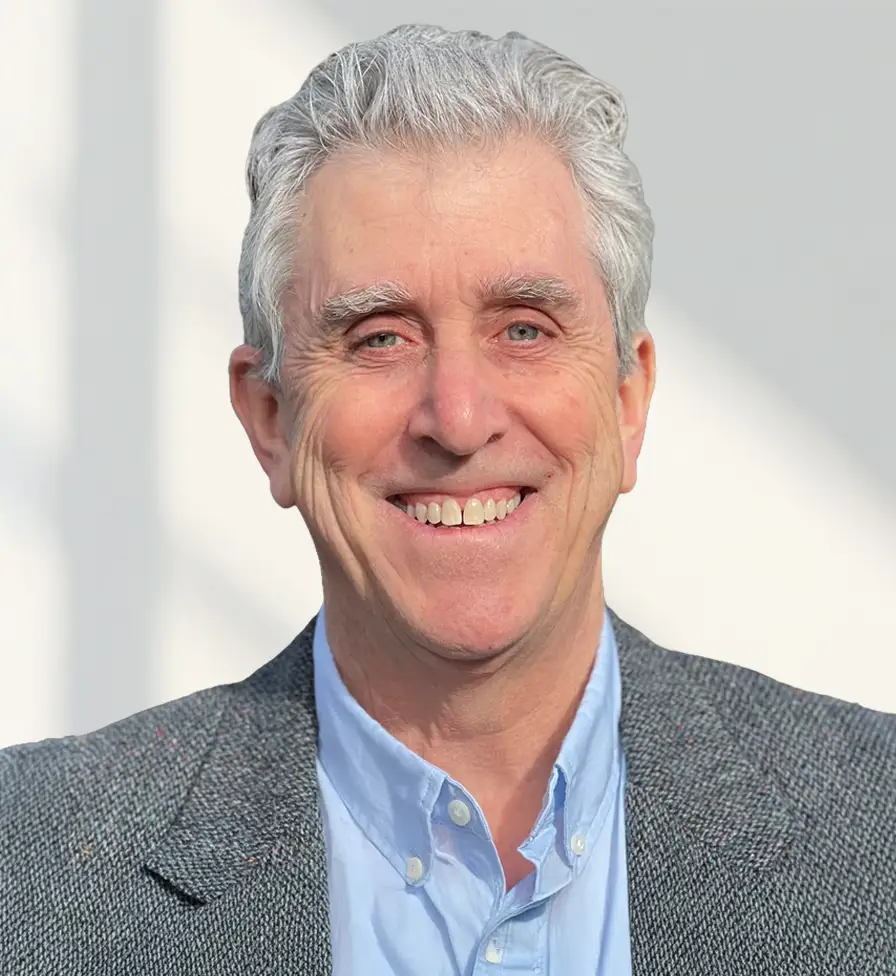 TraumaSolutions Smiling older man with gray hair wearing a gray blazer and light blue button-down shirt, standing in front of a plain light background, embodying the approachable demeanor of an experienced trauma therapist.