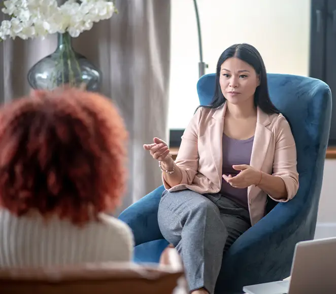TraumaSolutions A trauma therapist sits in a blue chair, speaking with another person whose back is to the camera, in a well-lit room with a large vase of white flowers in the background.