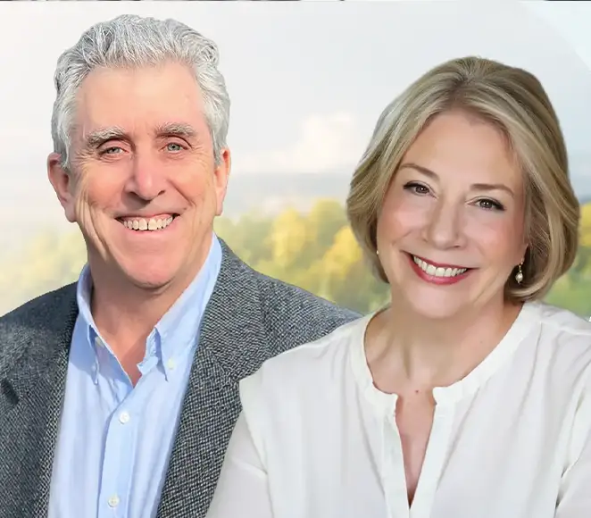 TraumaSolutions Two smiling older adults, one man in a blue shirt and gray blazer, and one woman in a white blouse, pose together in front of a blurred outdoor background, embodying the warm connection fostered by an experienced trauma therapist or compassionate EFT guide.