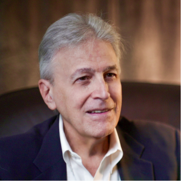 TraumaSolutions An older man with gray hair, wearing a dark blazer and white shirt, sits and smiles in a softly lit indoor setting.