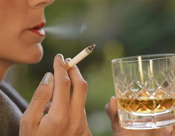 TraumaSolutions A person holding a lit cigarette in one hand and a glass of amber-colored liquid, likely alcohol, highlights the connection between smoking and drinking habits.