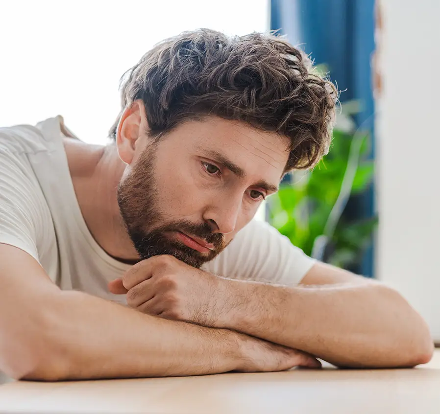 TraumaSolutions Man with curly hair and beard resting his chin on his arm, looking down with a sad or pensive expression, as if reflecting on a difficult connection or struggling with attachment.