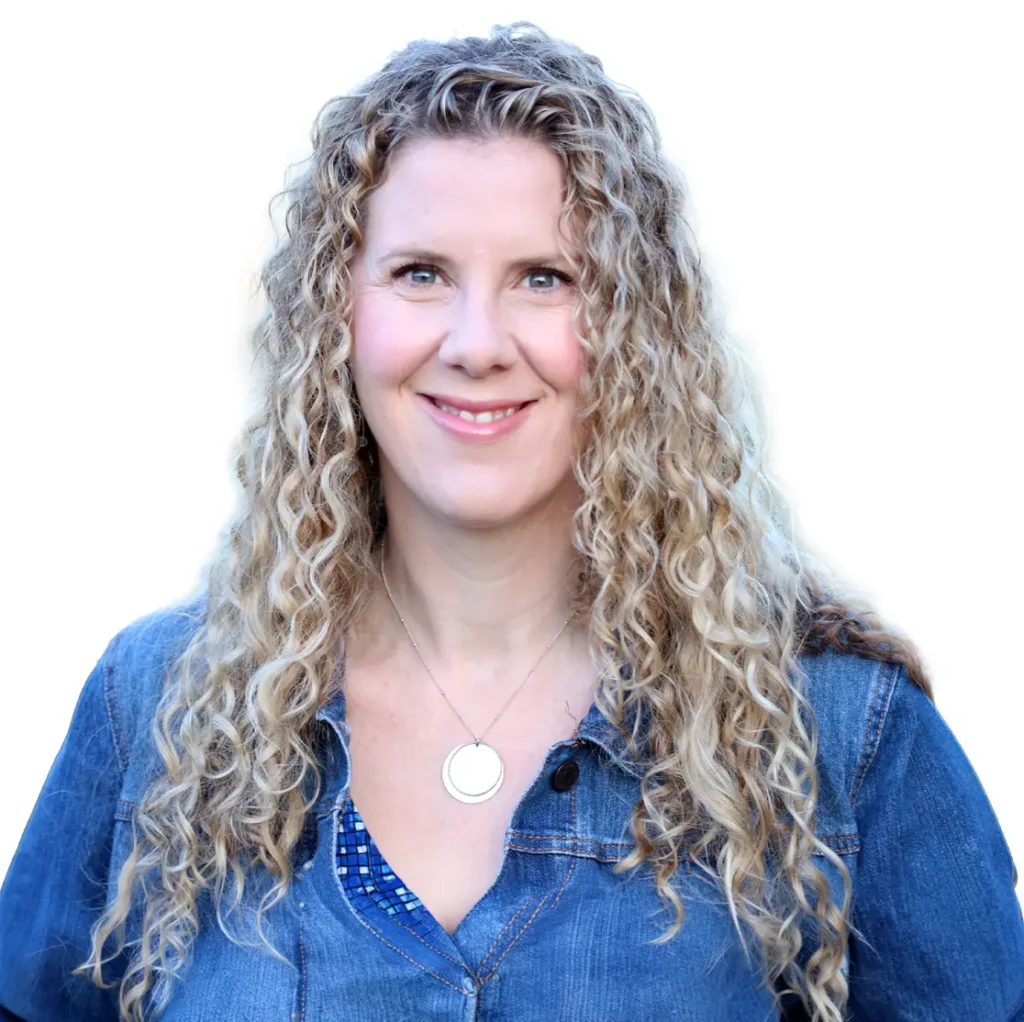 TraumaSolutions Woman with long curly blonde hair wearing a denim jacket and a circular pendant necklace, smiling in front of a plain white background, radiating warmth and connection.