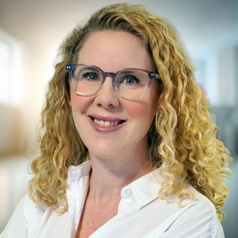 TraumaSolutions A woman with curly blonde hair, wearing glasses and a white button-up shirt, smiles in a brightly lit indoor setting, her expression reflecting warmth and genuine connection.