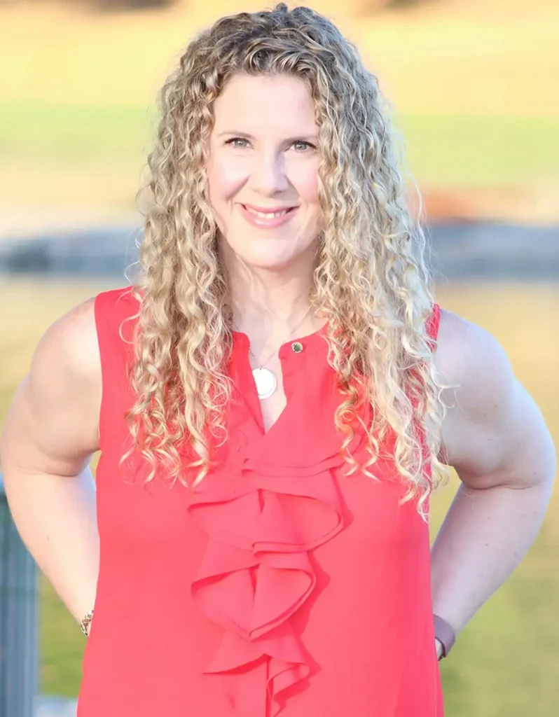 TraumaSolutions Woman with long, curly blonde hair wearing a sleeveless red blouse smiles outdoors in natural light, radiating warmth and connection against a softly blurred background.