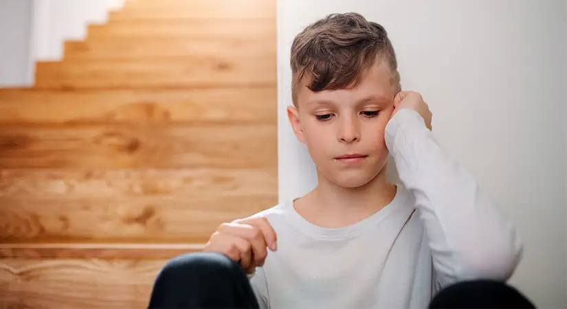 TraumaSolutions A boy in a white shirt sits at the bottom of wooden stairs, resting his head on his hand and looking down with a thoughtful expression, reflecting on the power of attachment and how we are all wired for connection.