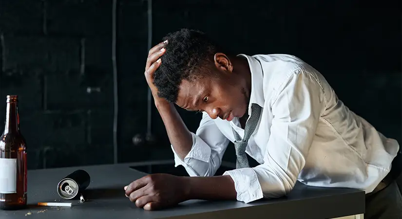 TraumaSolutions A man in a white shirt and loosened tie sits at a table, looking down with his head in one hand. An empty bottle and a can on the table hint at addiction, as he struggles with his thoughts in the quiet room.