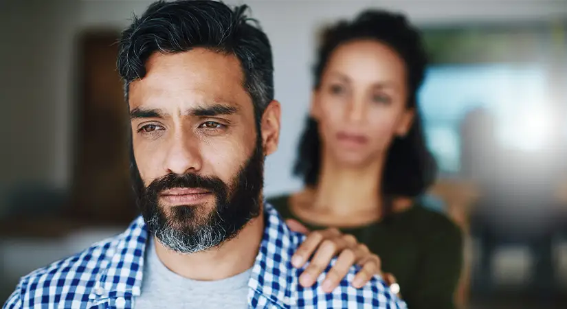TraumaSolutions A man looks pensive in the foreground while a woman stands behind him with her hand on his shoulder, offering support and fostering connection during a difficult moment.