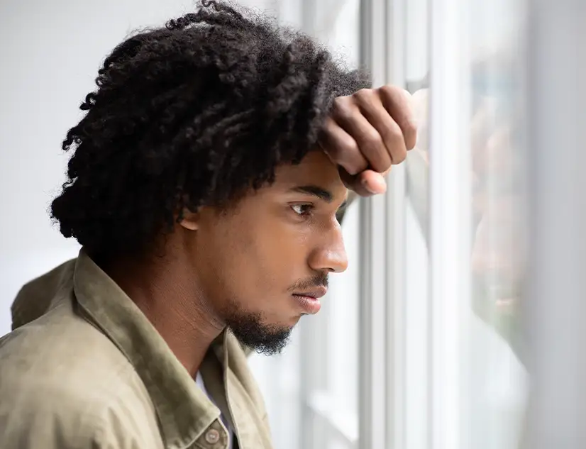TraumaSolutions A man with curly hair leans his forehead on his arm while looking out a window with a serious expression, reflecting on the complexity of attachment.