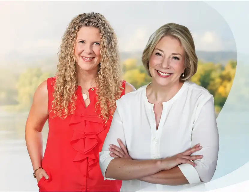 TraumaSolutions Two women standing side by side, one in a red sleeveless top with curly hair, the other in a white blouse with straight blonde hair, both smiling at the camera and showing their close connection.